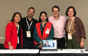 A group of panelists posing after a conference session, including one panelist participating via laptop