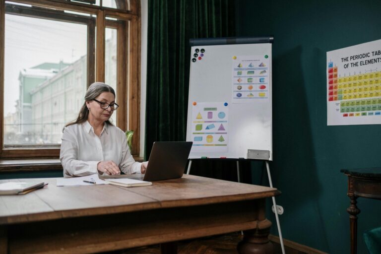 Female professor using a laptop while seated at a table in a classroom