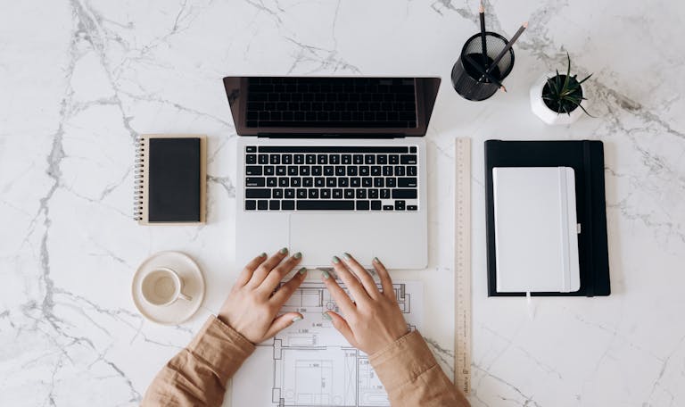 Top view of a stylish home office desk with a laptop, planner, and coffee cup, showing hands on a blueprint.