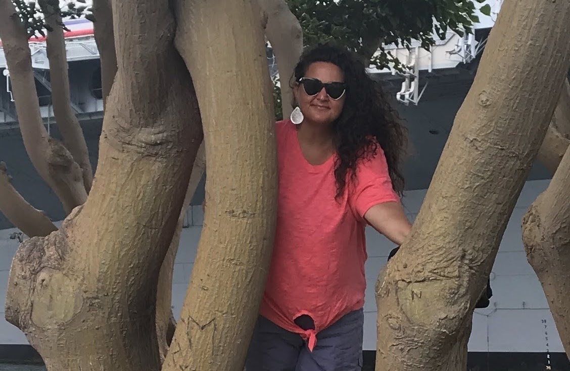 Woman wearing sunglasses and a bright pink shirt leaning up against a tree